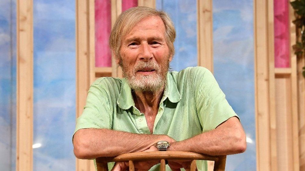Der deutsche Schauspieler Horst Janson lehnt lächelnd auf der Rückenlehne eines Stuhls. Er trägt ein hellgrünes Kurzarmhemd und eine silberne Armbanduhr. Im Hintergrund ist eine bunte Theaterkulisse mit Holzelementen und blauen sowie rosa Fensterausschnitten zu sehen.