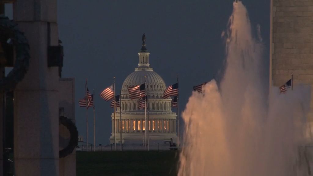 "Das Kapitol der Vereinigten Staaten in Washington, D.C., beleuchtet bei Nacht, umgeben von wehenden US-Flaggen und einem beleuchteten Springbrunnen im Vordergrund."